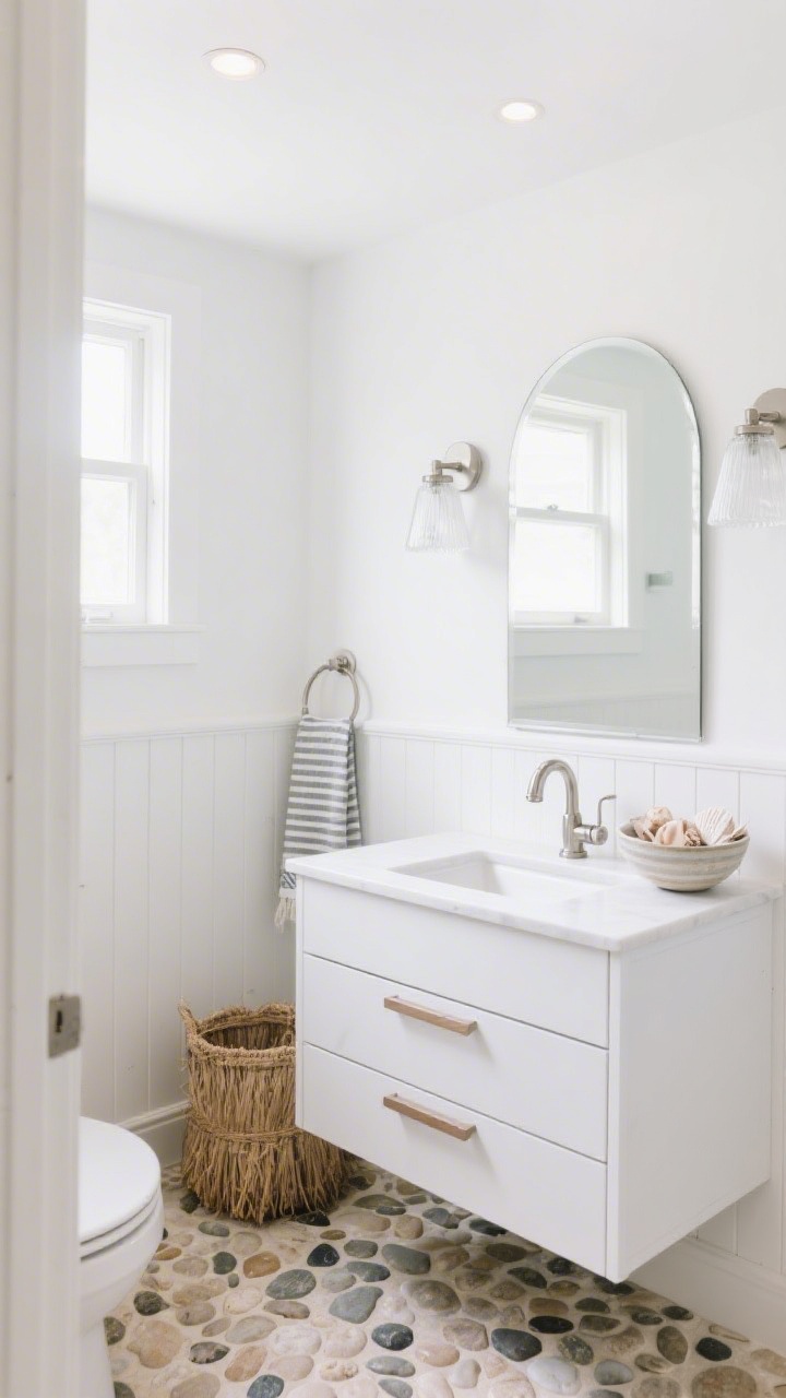 Wide shot: A small, windowless coastal-style bathroom in an all-over soft white palette, captured from a corner angle to show matte white shiplap on the lower half of the walls, a pebble-textured porcelain tile floor like beach pebbles, and a white oak floating vanity with slim drawers. An arched mirror above the sink reflects light from frosted glass sconces on either side; brushed nickel hardware and a high-arc faucet gleam softly. Style details include a striped Turkish towel on a brushed nickel hook, a sea grass hamper near the vanity, and a ceramic bowl of shells on the counter. Soft white, pale sand, and brushed nickel tones; recessed ceiling spots add ambient light. Photorealistic, bright and airy.